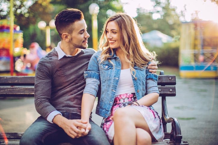 Young couple embracing and kissing in amusement park. They are sitting on the bench and enjoying in summer day.
