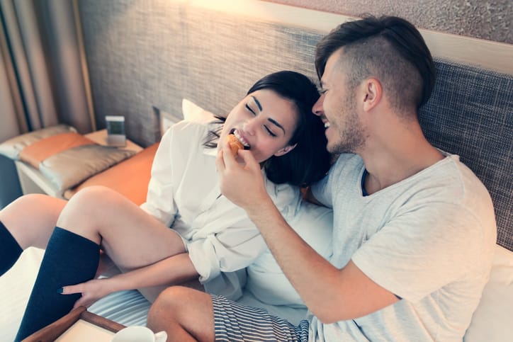 'Breakfast in bed' concept. Beautiful young love couple lying in bed, enjoying a carefree weekend morning together. Man is feeding his wife with a croissant.