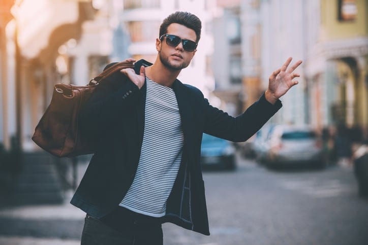 Handsome young man in smart casual wear carrying bag on shoulder and gesturing while hitchhiking on the street
