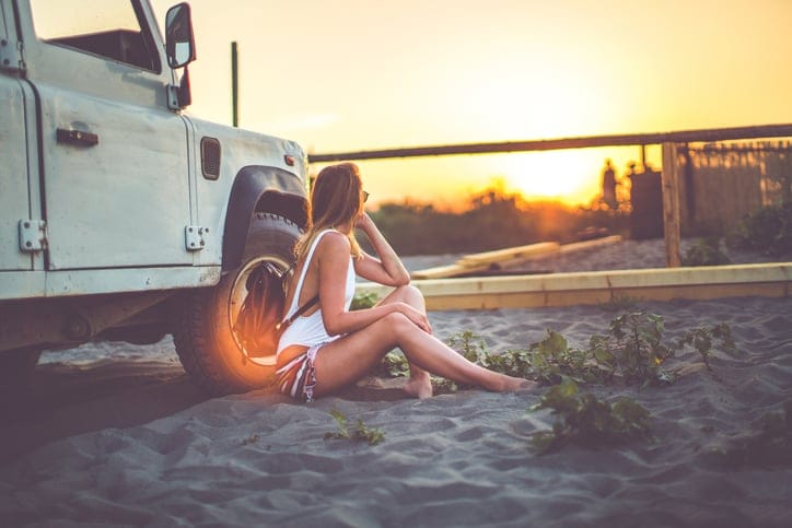 Girl sitting on the sandy beach next to a car and looking at the majestic sunset