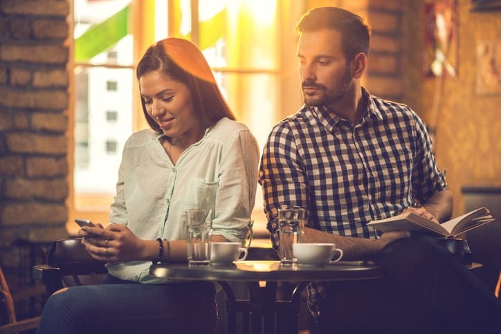 Smiling woman text messaging on cell phone in a cafe while her boyfriend is peeking to see to whom is she writing.