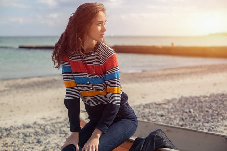 A girl sideways sitting near a red boat on the beach by the sea with beautiful sunset on the background. Woman in jeans, pullover and jacket on the sand