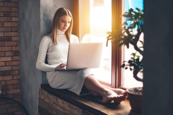 Beautiful young woman using her laptop while sitting at windowsill at home