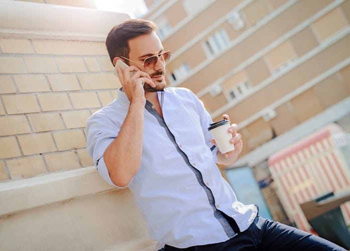 Handsome young smiling man standing on a street corner drinking coffee and talking on mobile phone. Lifestyle concept