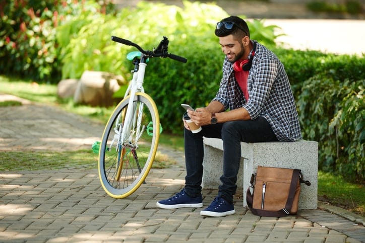 Smiling young man sitting on bench next to his bicycle and checking phone.