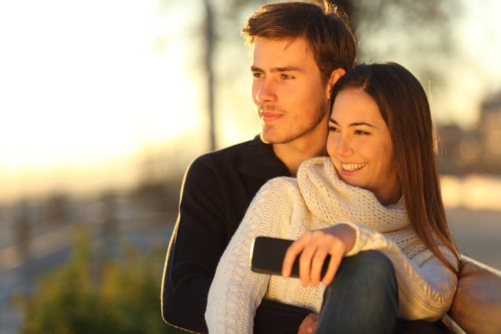Couple relaxing and hugging looking at sunset sitting on a bench and holding a smart phone