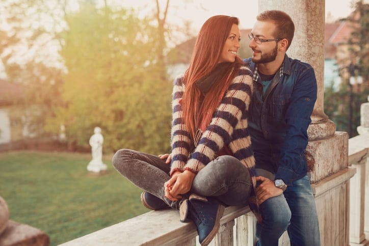 Young heterosexual couple sitting on the balcony fence, fondly looking at each other and smiling