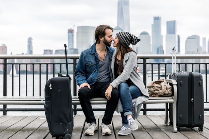 Young Couple Travelers Sitting on Bench and Waiting with Suitcases, bag with Lower Manhattan and the World Trade Center in the background.