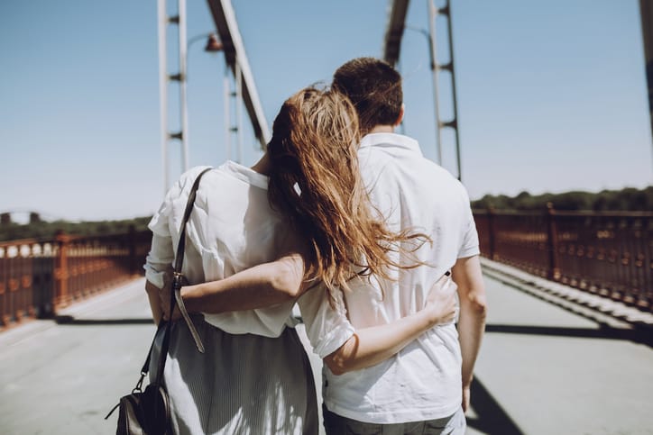 stylish couple in love hugging, back view with windy hair, on bridge in the summer city. modern woman and man in fashionable white clothes embracing at the river, romantic moment