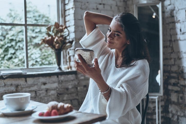 Young woman sitting at breakfast table, looking at herself in pocket mirror