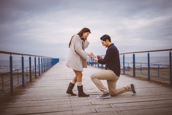 Man kneeling down proposing to his lover on a pier next to the sea
