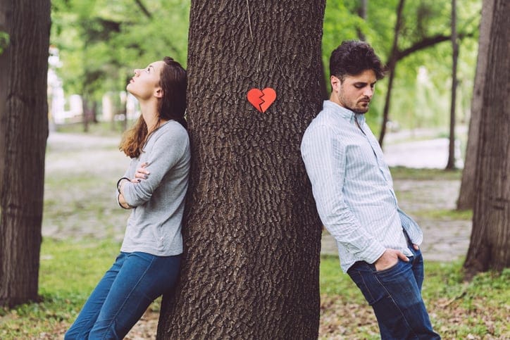 Unhappy couple leaning on opposite sides of tree trunk.