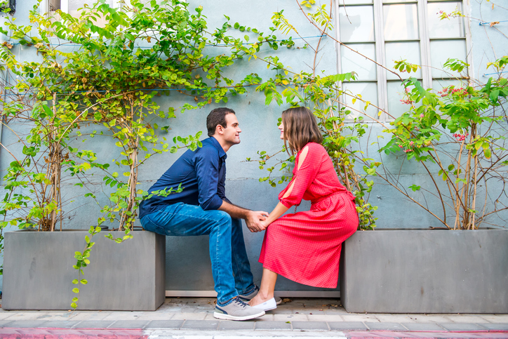 Profiles of young couple sitting opposite each other on the flower pots with green plants, holding hands and looking into eyes on the gray wall background. Love, dating, romance concept.