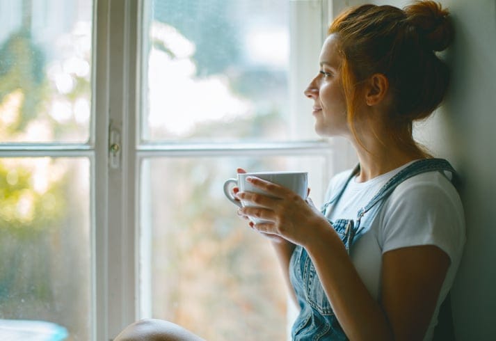 Young beautiful woman is looking through the window and drinking coffee in the morning