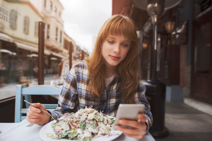 beautiful young woman is eating a salad in a restaurant while using an application in her smart phone