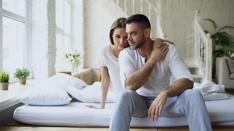 Depressed yong man sitting in bed having stressed while his girlfriend come and embrace him in bedroom at home