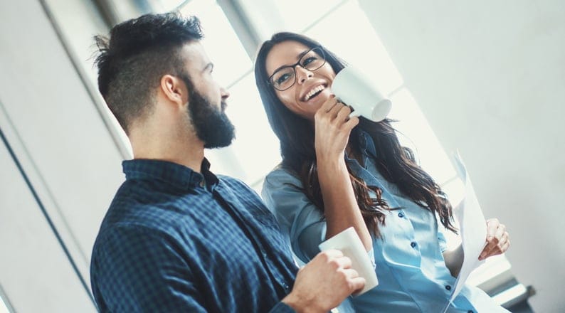 Closeup of two late 20's people taking a coffee break at their office. They are standing next to a window and having casual funny conversation. Tilt shot.