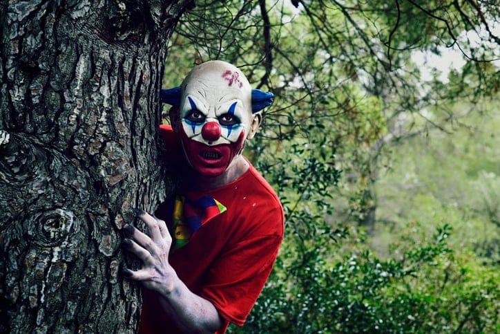 Scary evil clown beside a tree.