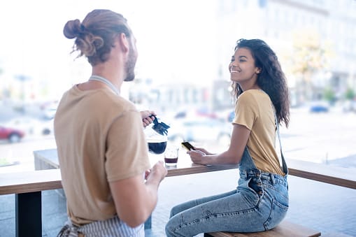 Time for coffee. Happy woman in denim overalls sitting in cafe near large window and man in apron with jug of coffee