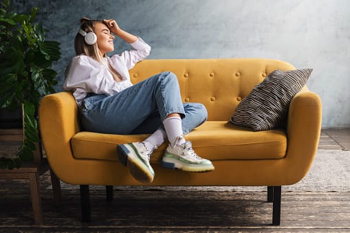 Young woman sitting on couch at home listening to music or podcast through headphones.