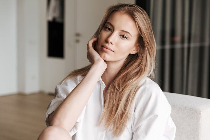 Beautiful young woman dressed in white shirt sitting on a couch at home and looking at camera