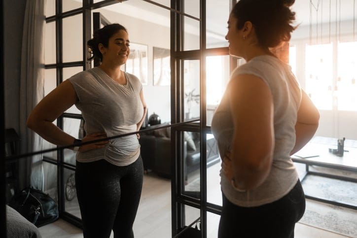 Woman doing some yoga exercises at home in her apartment. She is proudly looking at herself in the mirror with a smile.