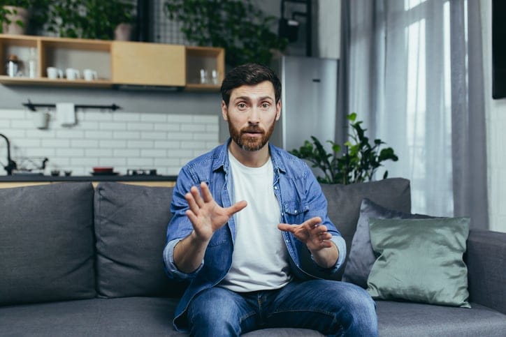 Concentrated man looking at webcam and talking on video call, sitting on sofa at home