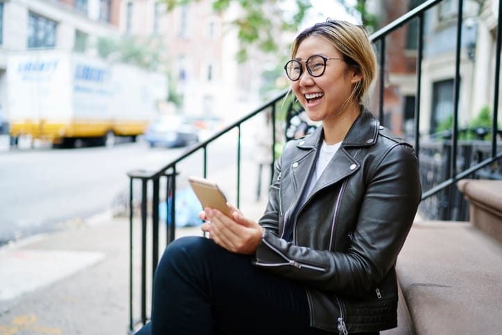 Cheerful Asian girl laughing at joke from social networks while sitting on stairs, happy Chinese female in glasses using application on smartphone for browsing phone mobility spending time in downtown