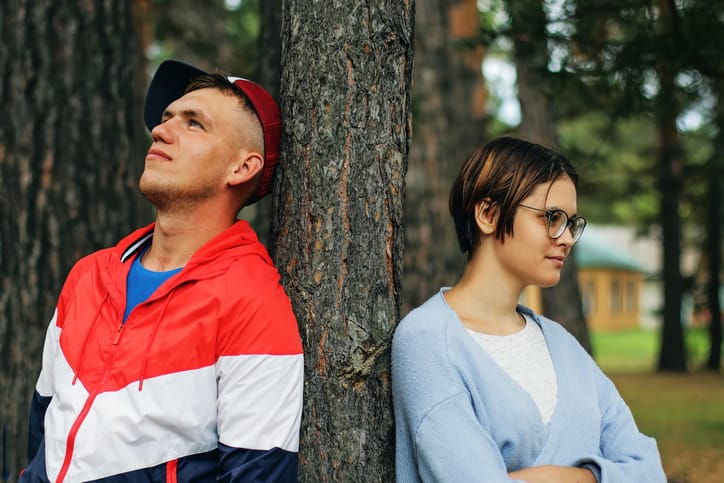 Two: a guy in a sport jacket and a baseball cap and a girl with a short haircut in glasses stand in nature near a tree after a quarrel, turning away from each other. The concept of misunderstanding and disagreement in relationships.