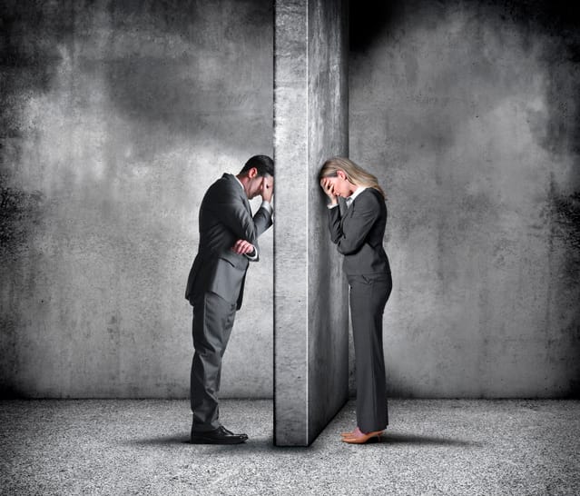 A businesswoman and a businessman place their heads in her hands as they lean in frustration against the wall that separates them.