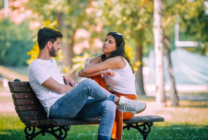 Happy casual couple talking looking each other sitting in a park with a green urban background