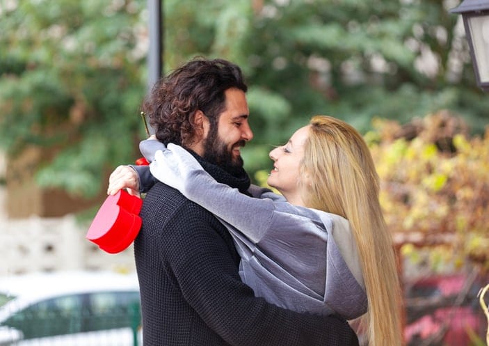 Young man hugging his girlfriend from behind and surprising her with a red rose.