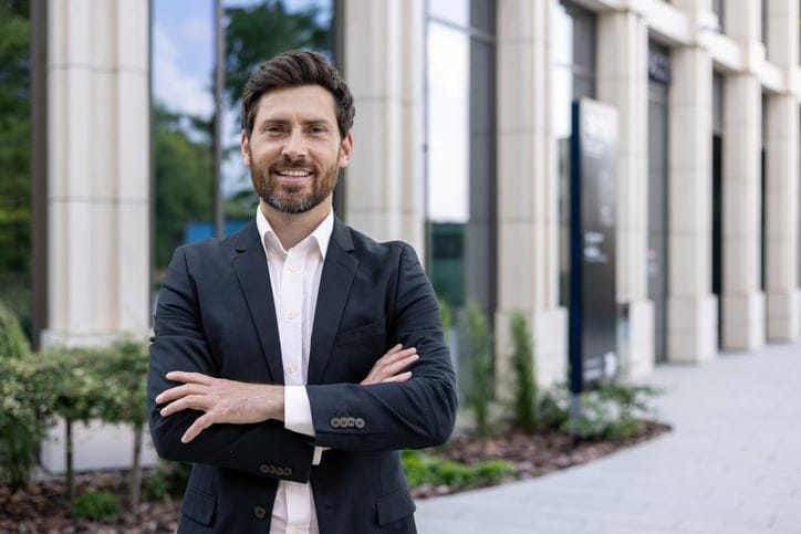 Portrait of a handsome young businessman, manager, leader standing in a suit near the office center and crossing his arms confidently smiling at the camera.
