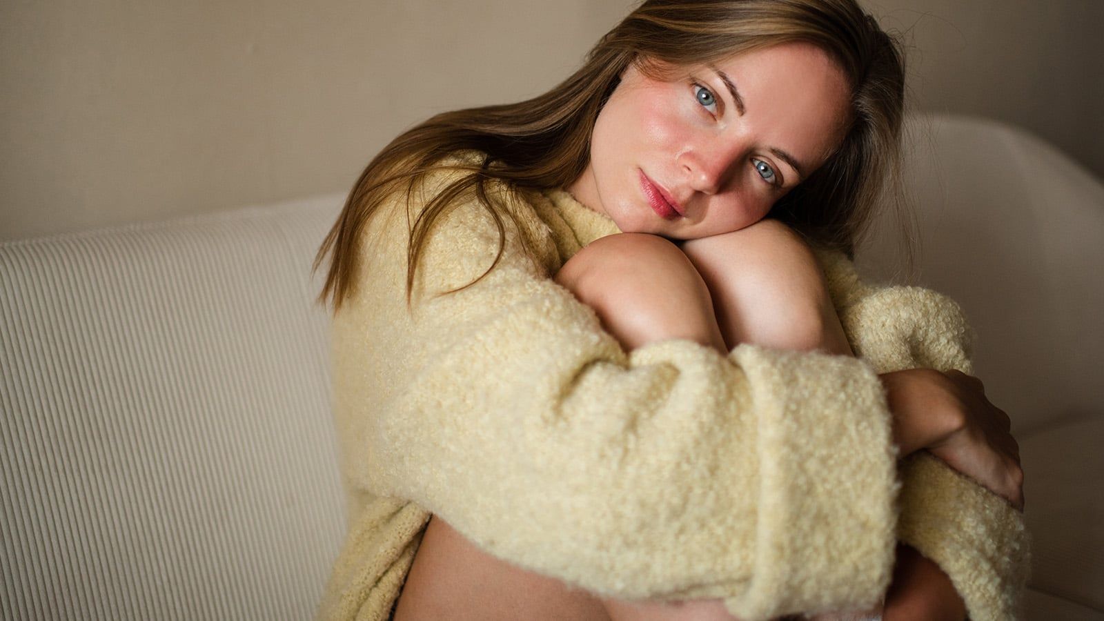 A woman with light brown hair wearing a soft, beige sweater sits on a white sofa, hugging her knees to her chest and looking at the camera with a gentle smile, radiating warmth and independence. The background is simple and neutral.