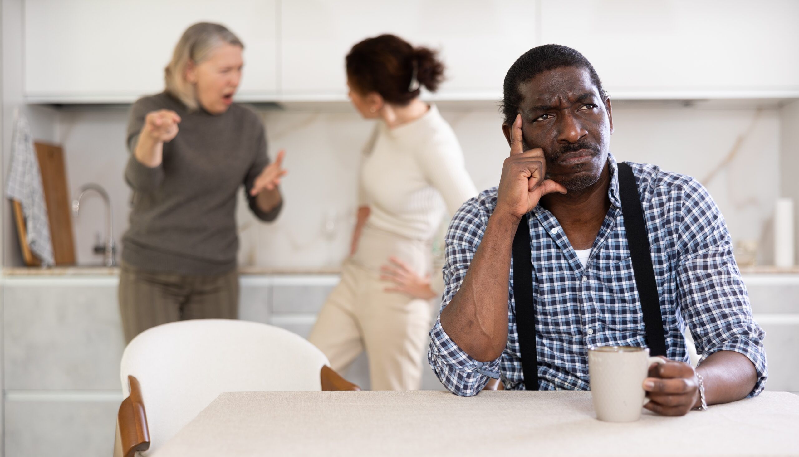 A sad middle aged man sitting at the kitchen table and not part of a conversation going on behind him