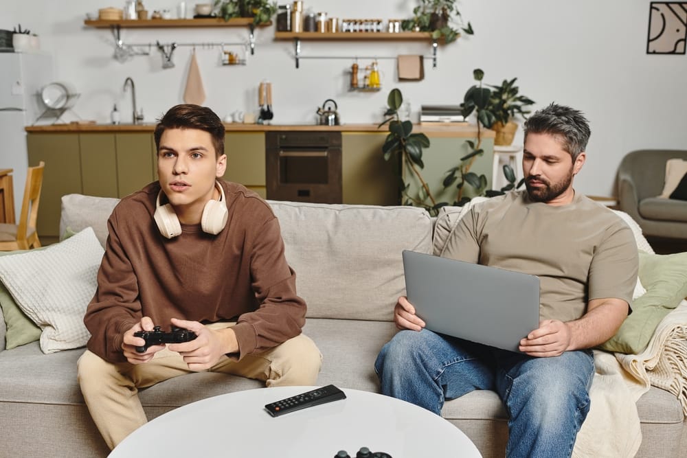 Two friends sitting on the couch with one using laptop and the other playing games