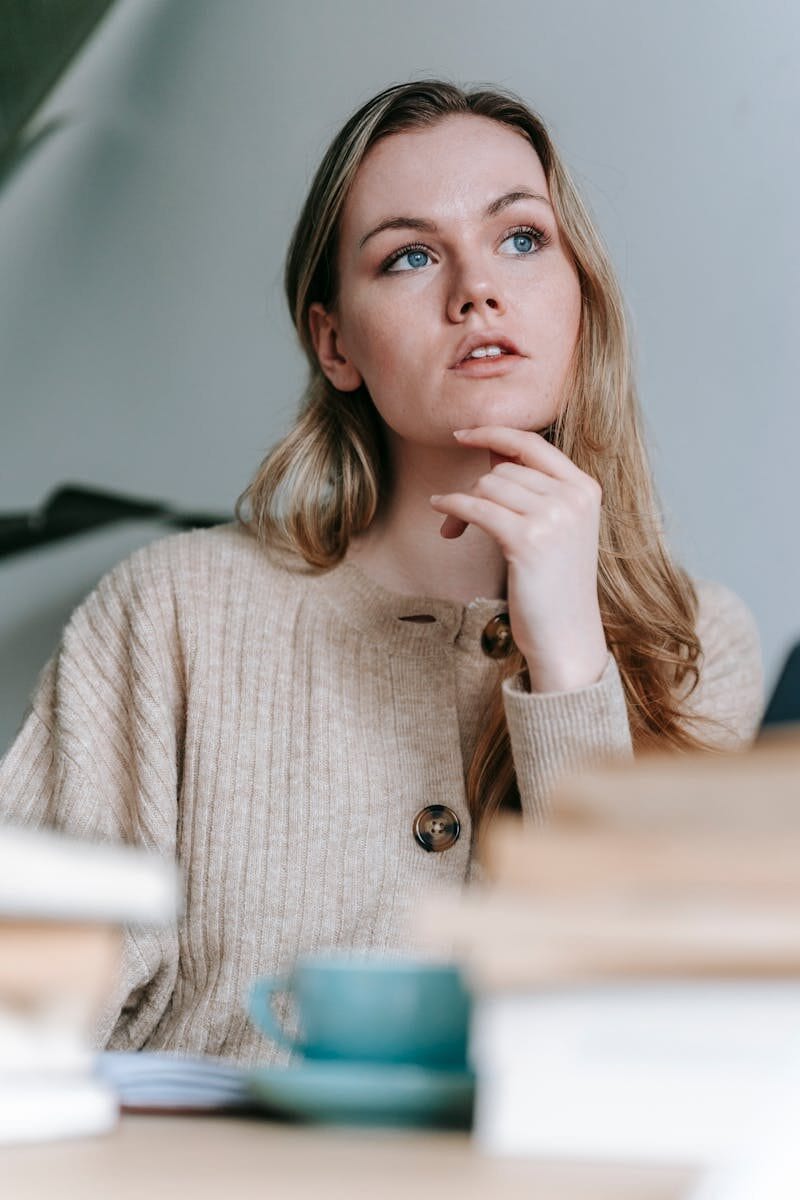 Thoughtful female with long hair touching chin and contemplating at table with cup and many books