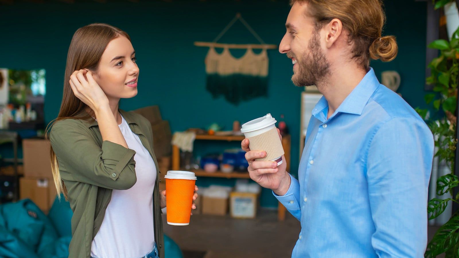 A woman and a man stand indoors, smiling and engaging in small talk while holding takeaway coffee cups. The woman touches her hair, and the man has his hair tied back. The background is a cozy, casual workspace with teal walls.