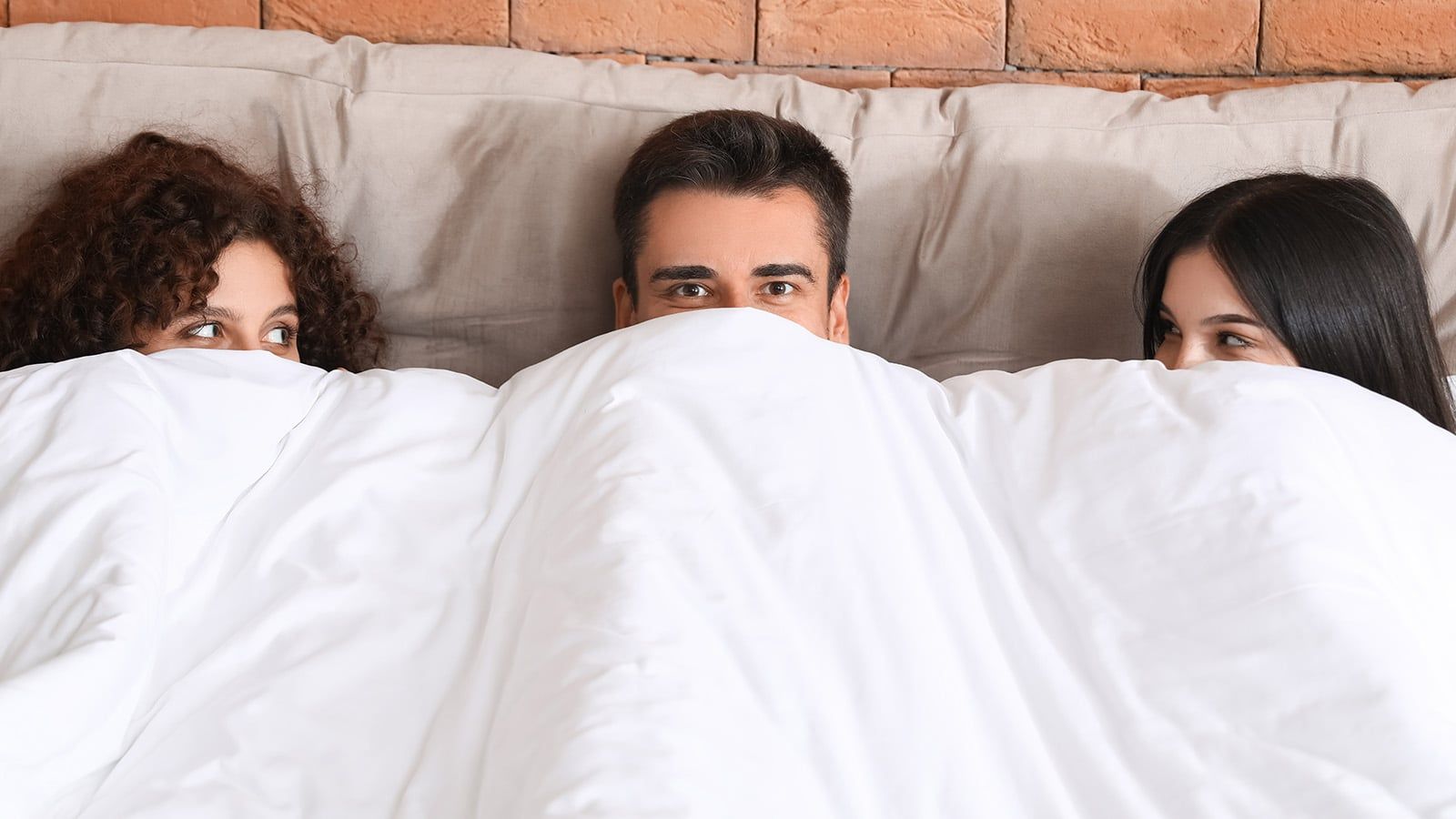 Three people lying in bed, partially covered by a white blanket, with only their faces visible and smiling at each other. They appear relaxed and cozy against a light gray headboard.