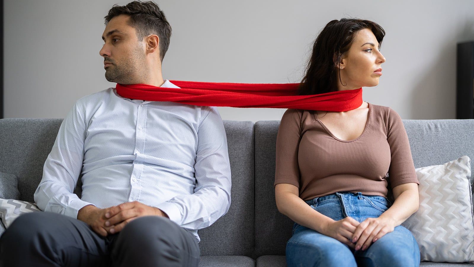 A man and woman sit apart on a couch, both looking away from each other, with a red scarf looped around both their necks, symbolizing tension or a strained relationship.