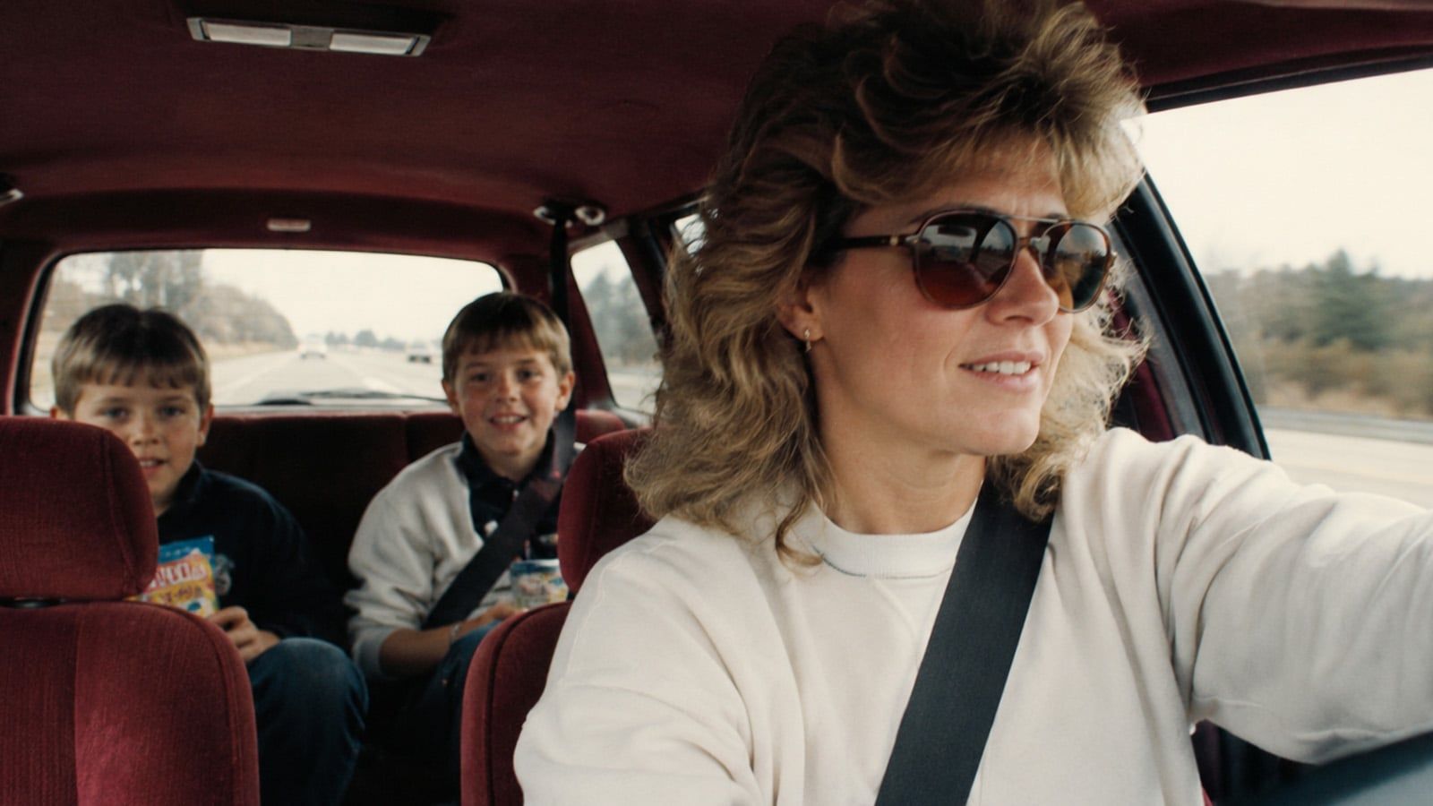 A woman with feathered hair and sunglasses drives a car, smiling, with two young boys in the back seat. The boys appear cheerful, holding snacks, and the scene is set on a highway during the day.