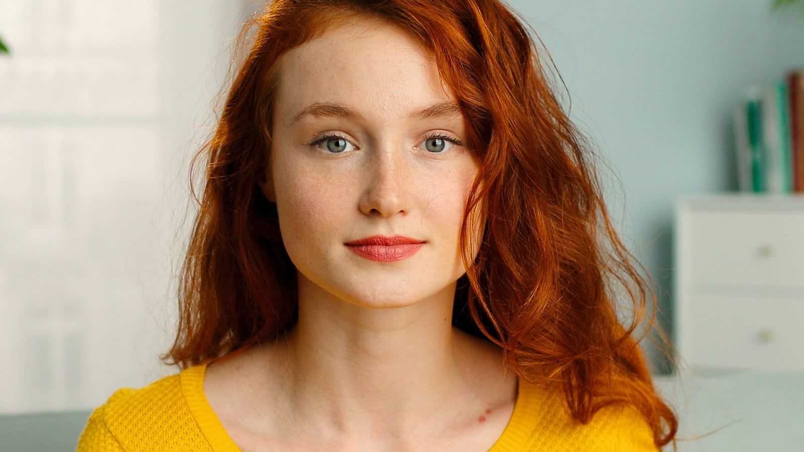 A young woman with long red hair and fair skin, wearing a yellow sweater, looks directly at the camera with a neutral expression. The background is softly blurred, showing light-colored walls and shelves.