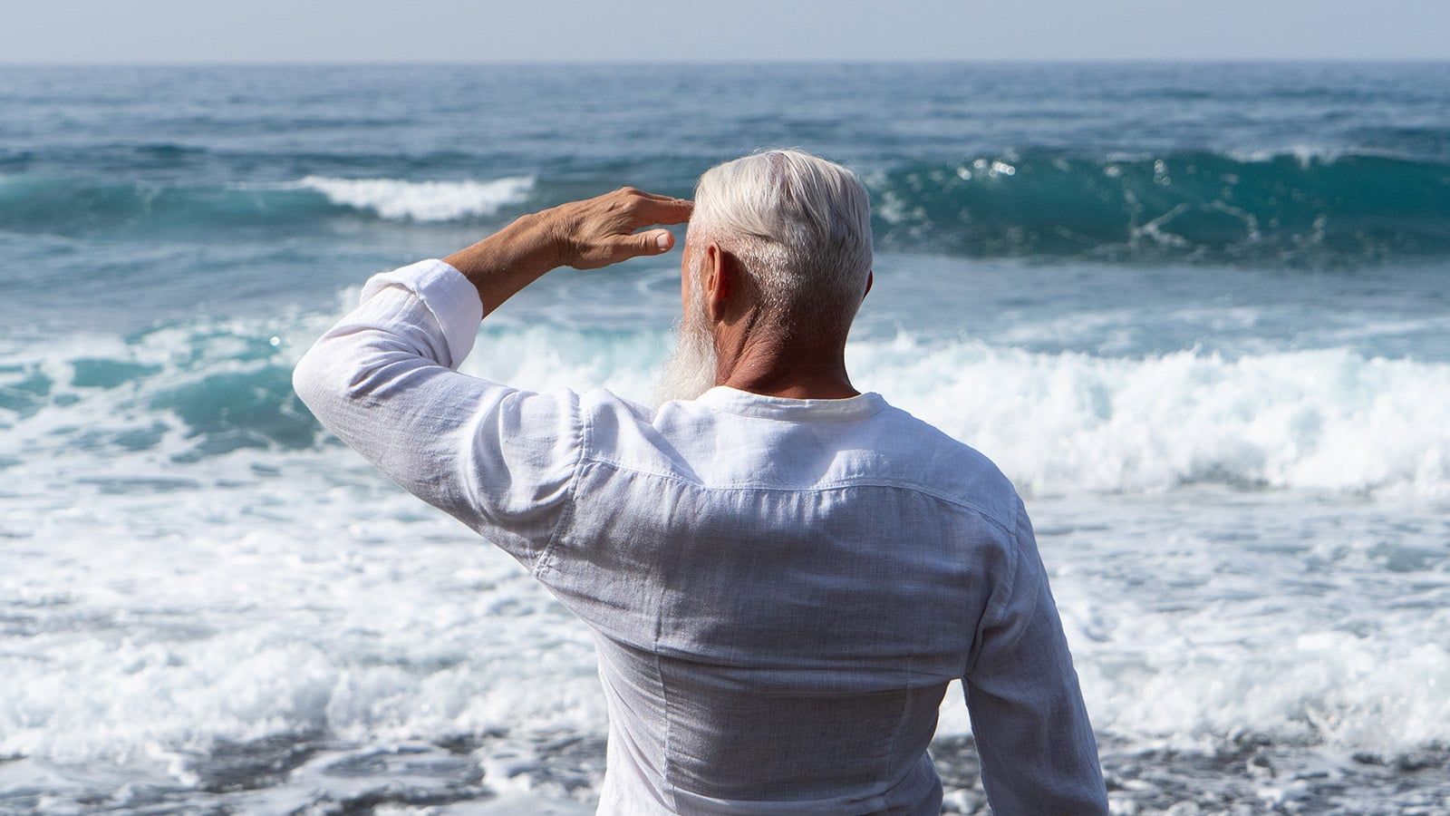 An older man with white hair and a beard stands on the shore, facing the ocean, saluting with his right hand. He wears a light-colored long-sleeve shirt, and waves are crashing in the background.