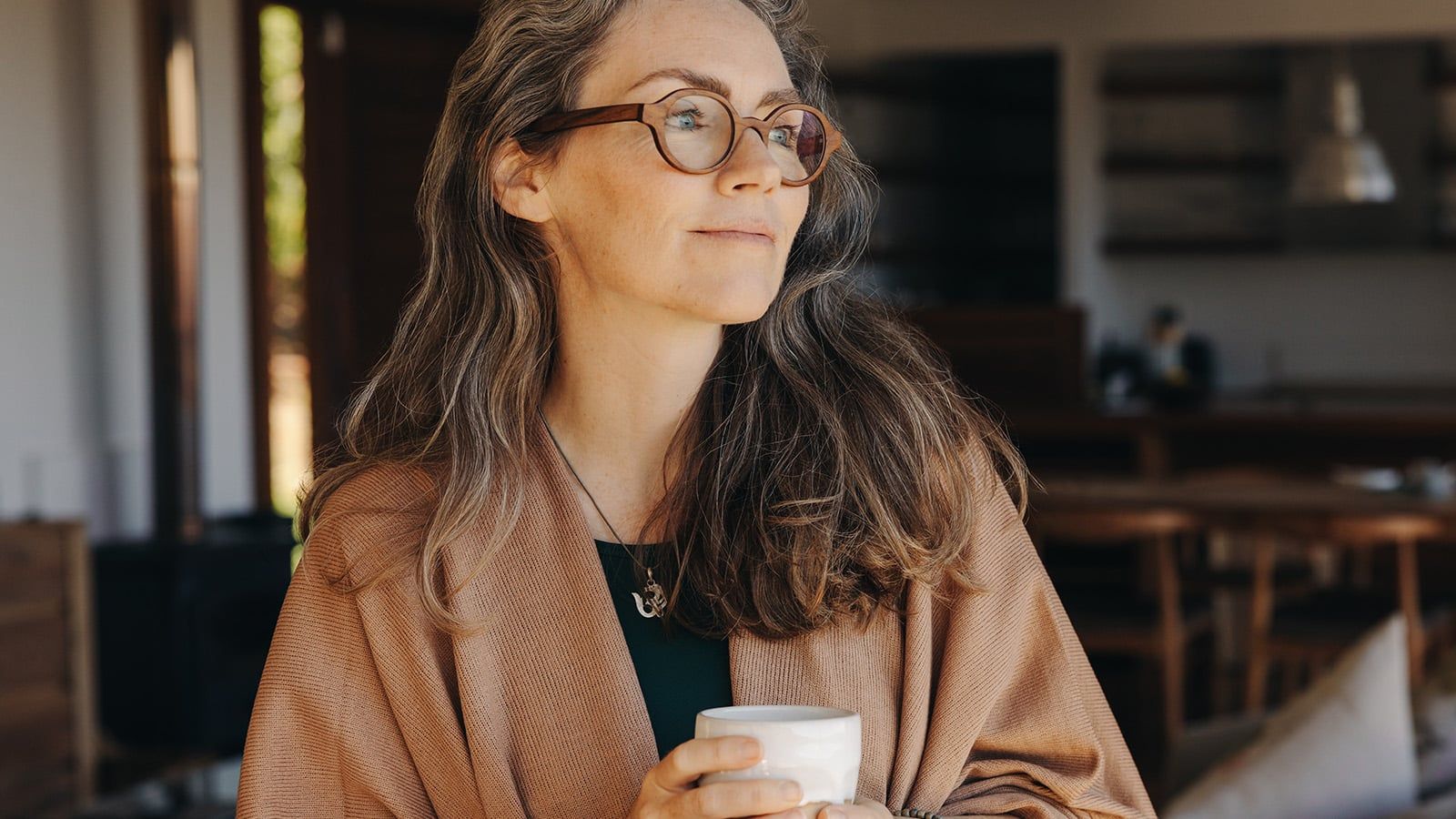 A woman with long, wavy hair and glasses holds a white mug, gazing thoughtfully to the side. Wrapped in a tan shawl, she stands in a cozy room with wooden furniture, reflecting on her successful retirement.