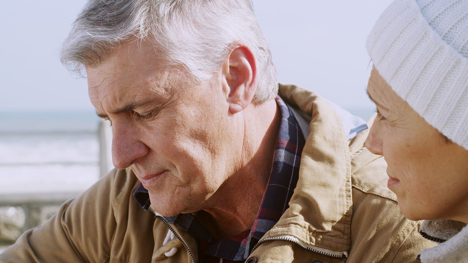 A close-up of an older man looking down thoughtfully, wearing a brown jacket and plaid shirt, with a woman in a white knit hat beside him. They are outdoors near the ocean.