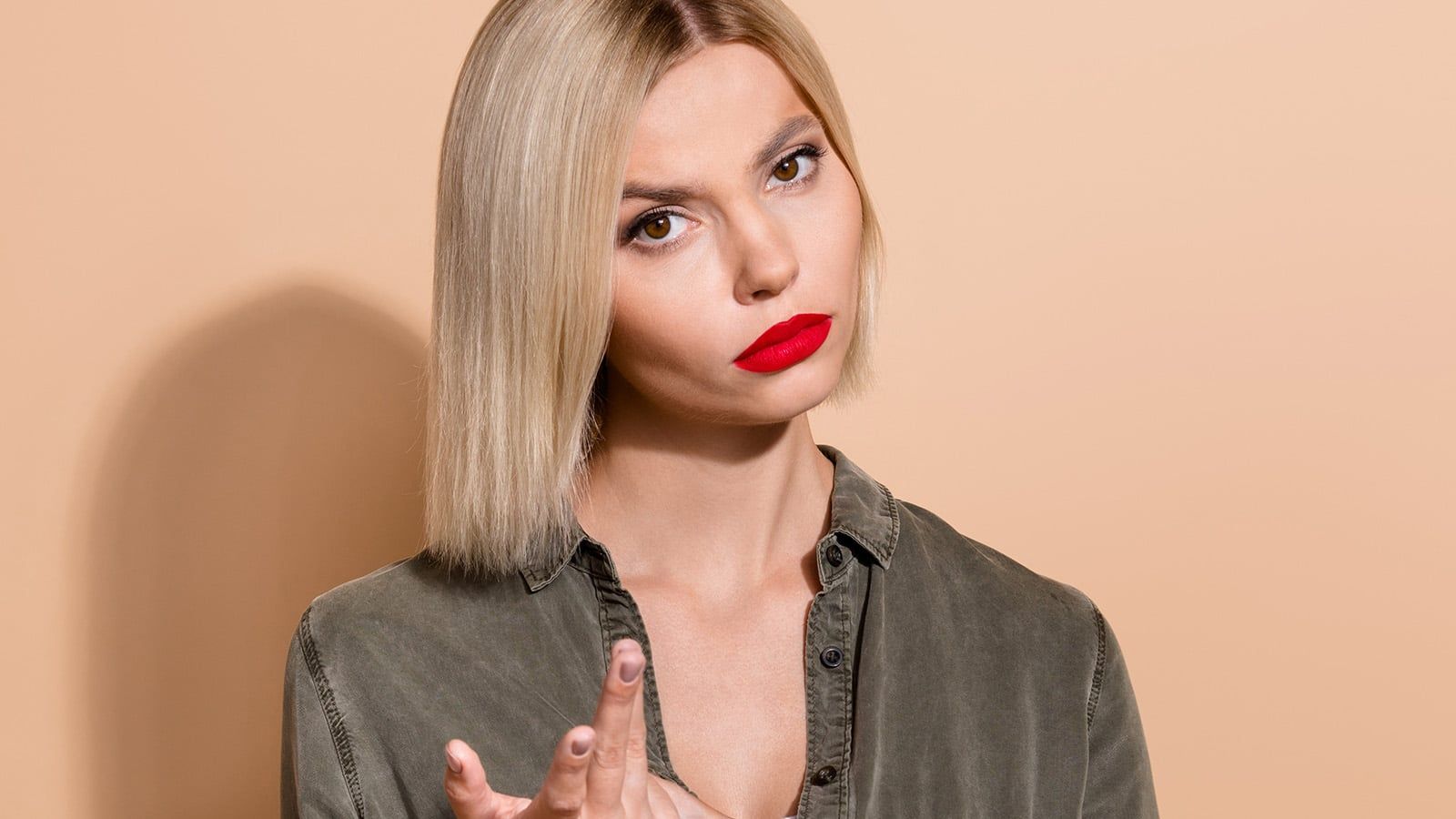 A woman with straight blonde hair and bright red lipstick looks seriously at the camera, wearing a green button-up shirt and holding up her hand with two fingers extended, as if challenging expectations, against a beige background.