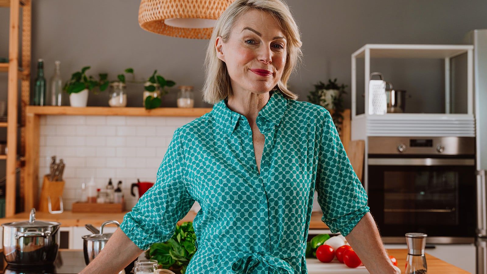 A smiling woman with short blonde hair wearing a teal patterned dress stands in a modern kitchen with wooden shelves, green plants, and fresh vegetables on the counter.