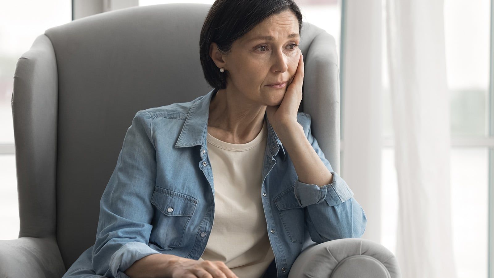 A woman with short dark hair sits in a gray armchair, resting her head on her hand and looking thoughtful or concerned. She wears a light denim shirt over a white top. Light streams in from a window behind her.