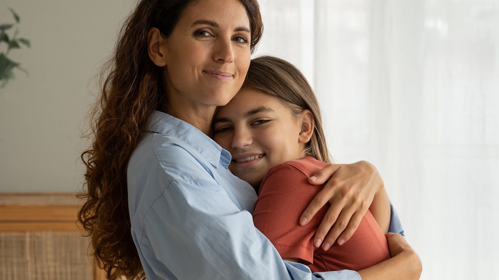 A woman with long, wavy hair lovingly hugs a smiling young girl in a warmly lit room with sheer curtains in the background. Both appear happy and comfortable.