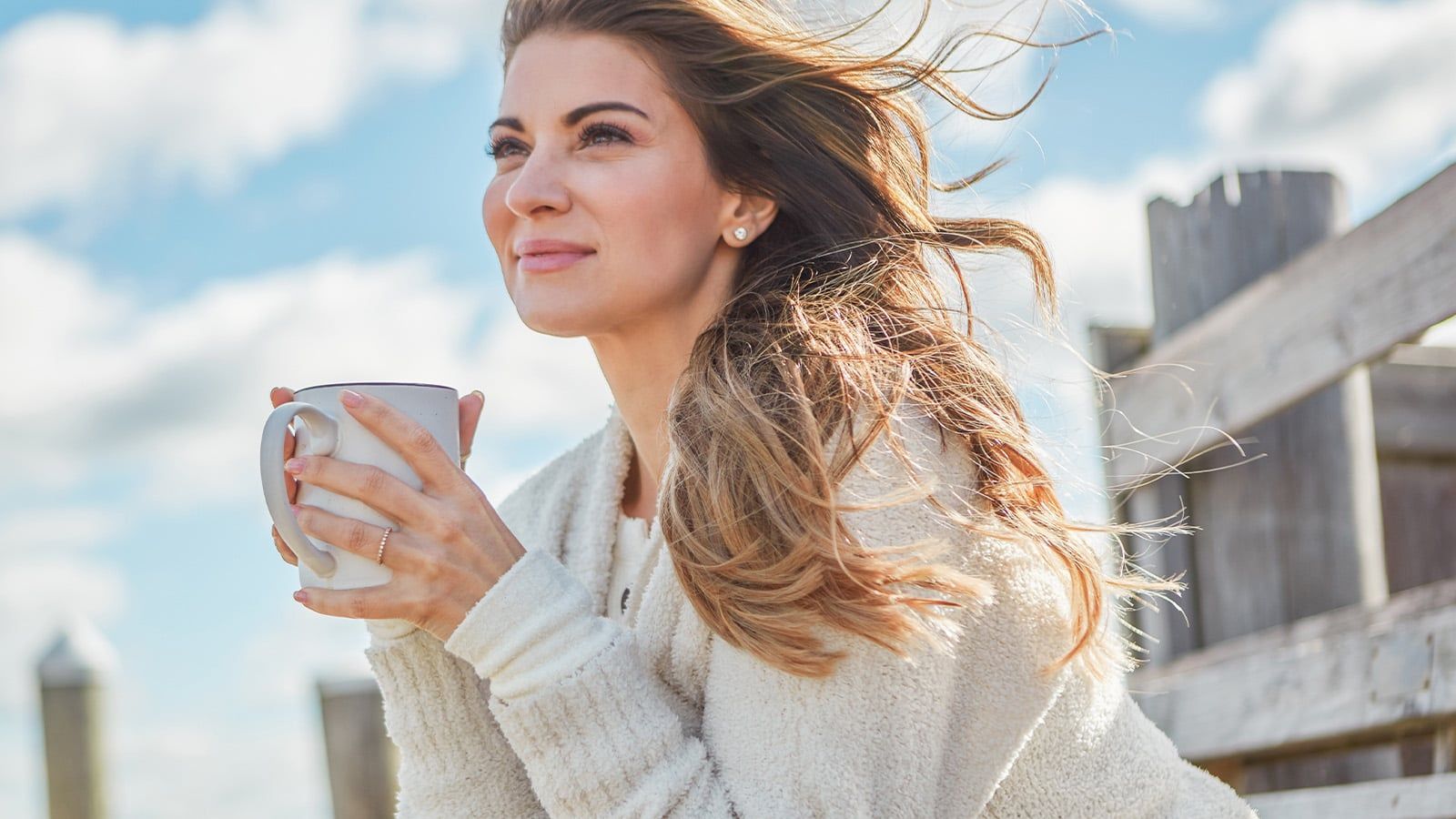 A woman with long hair sits outdoors on a sunny day, holding a mug and smiling. She wears a light-colored sweater, and the wind is blowing her hair. The sky is blue with clouds, and a wooden fence is in the background.